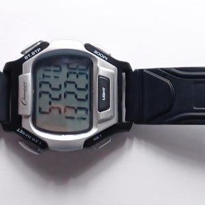 May include: A digital wristwatch with a silver-colored case and a black rubber strap. The watch face displays the time and date. The strap has a buckle closure and multiple adjustment holes. The watch is designed for everyday wear.
