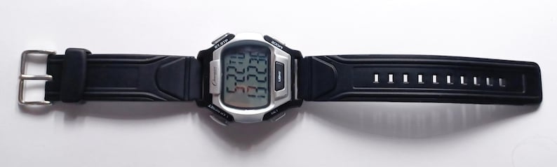 May include: A digital wristwatch with a silver-colored case and a black rubber strap. The watch face displays the time and date. The strap has a buckle closure and multiple adjustment holes. The watch is designed for everyday wear.