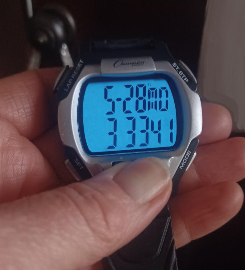 May include: A Champion digital watch with a black strap and silver bezel. The watch face displays the time as 5:28 and a four-digit number. The watch has buttons labeled LAP RESET, ST.STP, SET, and MODE.