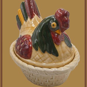 May include: A ceramic hen-shaped container with a basket-weave base. The hen has a red comb and wattle, green and red feathers, and a yellow beak. The container is cream-colored with a glossy finish.