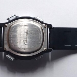 May include: Black digital watch with a rectangular face and a black rubber strap. The watch has a silver buckle and a silver back. The strap has a series of rectangular holes. The watch is lying flat on a white surface.