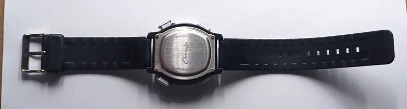 May include: Black digital watch with a rectangular face and a black rubber strap. The watch has a silver buckle and a silver back. The strap has a series of rectangular holes. The watch is lying flat on a white surface.