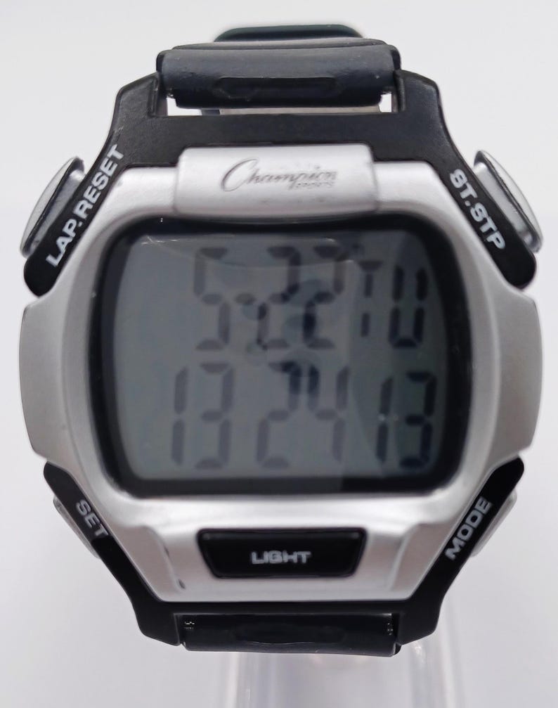 May include: A digital Champion watch with a silver face and black accents. The watch displays the time and date. It has buttons labeled LAP.RESET, ST.STP, SET, MODE, and LIGHT. The Champion logo is visible on the watch face.