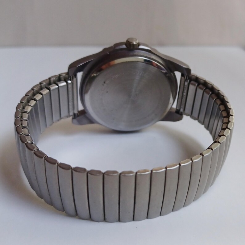 K&ouml;nnte beinhalten: Eine silberfarbene Armbanduhr mit einem dehnbaren Metallarmband. Das Zifferblatt der Uhr ist rund, und die R&uuml;ckseite der Uhr ist sichtbar. Das Armband besteht aus kleinen, rechteckigen Metallgliedern, die sich dehnen.