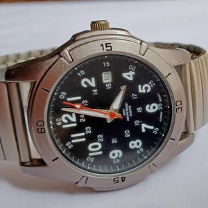 K&ouml;nnte beinhalten: Eine silberfarbene Armbanduhr mit schwarzem Zifferblatt, wei&szlig;en Ziffern und einem roten Sekundenzeiger. Die Uhr hat eine Datumsanzeige und ein flexibles Metallarmband. Auf dem Zifferblatt steht "Japan 30 M". Der Durchmesser betr&auml;gt ca. 4 cm.
