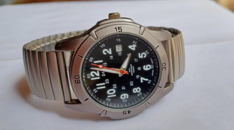 K&ouml;nnte beinhalten: Eine silberfarbene Armbanduhr mit schwarzem Zifferblatt, wei&szlig;en Ziffern und einem roten Sekundenzeiger. Die Uhr hat eine Datumsanzeige und ein flexibles Metallarmband. Auf dem Zifferblatt steht "Japan 30 M". Der Durchmesser betr&auml;gt ca. 4 cm.