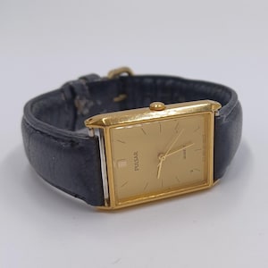 May include: A gold-toned rectangular Pulsar Quartz watch with a black leather band. The watch face is gold with black hour markers and hands. The watch is a classic design.