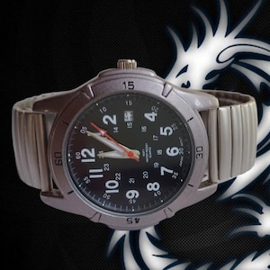 K&ouml;nnte beinhalten: Silberfarbene Armbanduhr mit schwarzem Zifferblatt und wei&szlig;en Ziffern. Datumsanzeige bei 3 Uhr und roter Sekundenzeiger. Das Armband ist silberfarben und dehnbar. Auf der Uhr steht 'Japan 3D Movt'.