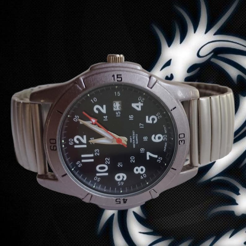 K&ouml;nnte beinhalten: Silberfarbene Armbanduhr mit schwarzem Zifferblatt und wei&szlig;en Ziffern. Datumsanzeige bei 3 Uhr und roter Sekundenzeiger. Das Armband ist silberfarben und dehnbar. Auf der Uhr steht 'Japan 3D Movt'.