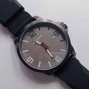 K&ouml;nnte beinhalten: Eine Armbanduhr mit einem runden, dunkelmetallischen Geh&auml;use und einem glatten schwarzen Silikonarmband. Das Zifferblatt ist in einem gedeckten Braun gehalten und zeigt gro&szlig;e wei&szlig;e arabische Ziffern bei 12, 3, 6 und 9, wei&szlig;e Indizes, wei&szlig;e Zeiger und den Schriftzug ROUSSEAU.