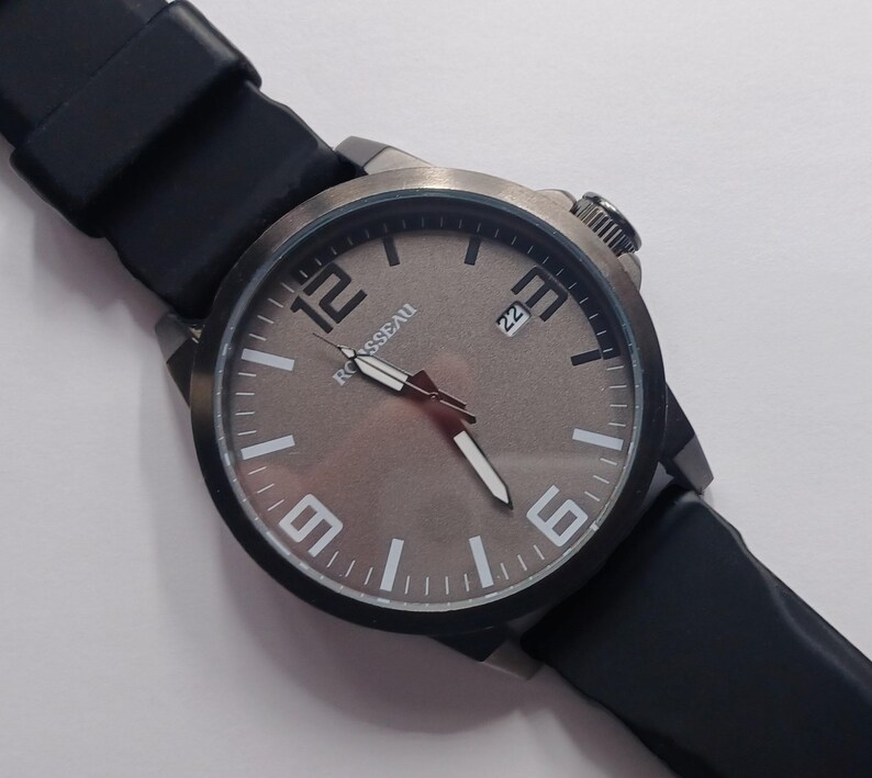 K&ouml;nnte beinhalten: Eine Armbanduhr mit einem runden, dunkelmetallischen Geh&auml;use und einem glatten schwarzen Silikonarmband. Das Zifferblatt ist in einem gedeckten Braun gehalten und zeigt gro&szlig;e wei&szlig;e arabische Ziffern bei 12, 3, 6 und 9, wei&szlig;e Indizes, wei&szlig;e Zeiger und den Schriftzug ROUSSEAU.
