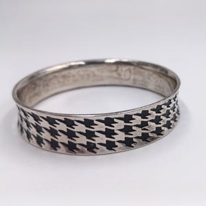 May include: A silver-toned bangle bracelet with a black and white houndstooth pattern around the band. The bracelet is circular and appears to be made of metal. The background is plain white.