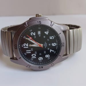 K&ouml;nnte beinhalten: Eine silberfarbene Armbanduhr mit schwarzem Zifferblatt und silbernem, dehnbarem Armband. Das Zifferblatt zeigt wei&szlig;e Ziffern, rote und wei&szlig;e Zeiger und den Text "WATER RESIST QUARTZ JAPAN 30 MONT". Das Armband ist flexibel.