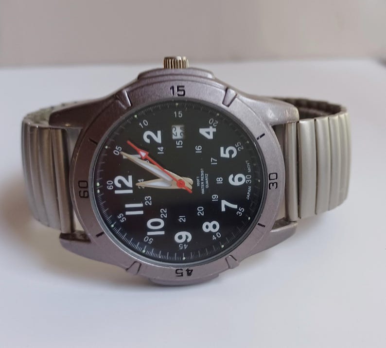 K&ouml;nnte beinhalten: Eine silberfarbene Armbanduhr mit schwarzem Zifferblatt und silbernem, dehnbarem Armband. Das Zifferblatt zeigt wei&szlig;e Ziffern, rote und wei&szlig;e Zeiger und den Text "WATER RESIST QUARTZ JAPAN 30 MONT". Das Armband ist flexibel.