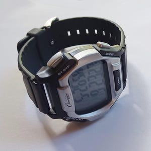 May include: A digital Champion watch with a black rubber strap and a silver-colored face. The watch displays the time on a digital screen and has buttons for mode, light, lap reset, and start/stop. The watch is on a white surface.