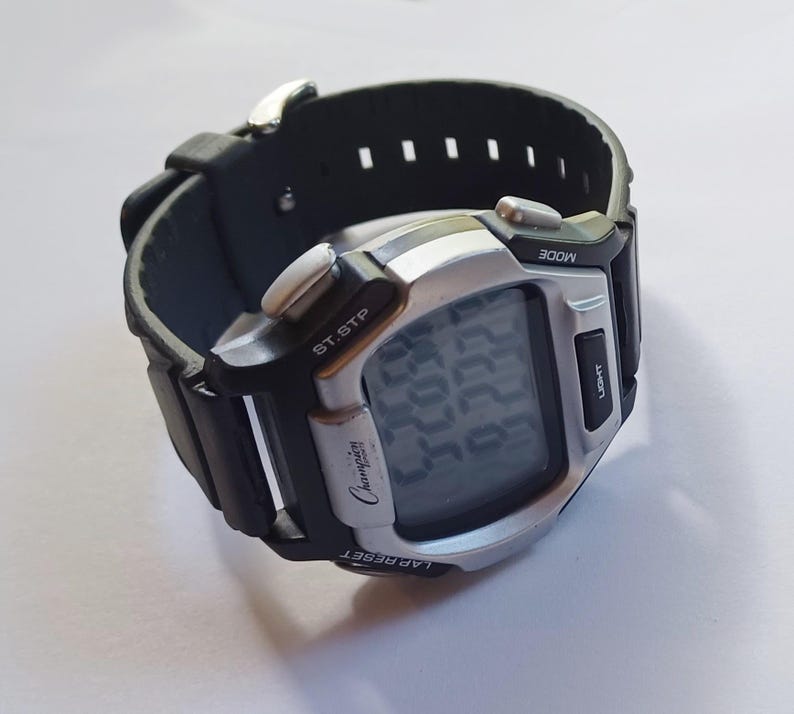 May include: A digital Champion watch with a black rubber strap and a silver-colored face. The watch displays the time on a digital screen and has buttons for mode, light, lap reset, and start/stop. The watch is on a white surface.
