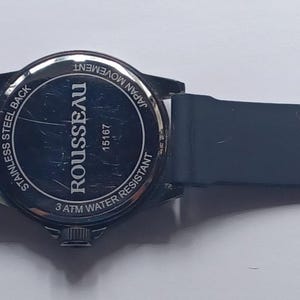 K&ouml;nnte beinhalten: Eine schwarze Armbanduhr mit Silikonarmband und Geh&auml;useboden aus Edelstahl. Auf der R&uuml;ckseite der Uhr ist der Text ROUSSEAU, 18167, JAPAN MOVEMENT, STAINLESS STEEL BACK und 2 ATM WATER RESISTANT in einem kreisf&ouml;rmigen Muster aufgedruckt.