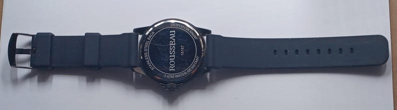 K&ouml;nnte beinhalten: Eine schwarze Armbanduhr mit Silikonarmband und Geh&auml;useboden aus Edelstahl. Auf der R&uuml;ckseite der Uhr ist der Text ROUSSEAU, 18167, JAPAN MOVEMENT, STAINLESS STEEL BACK und 2 ATM WATER RESISTANT in einem kreisf&ouml;rmigen Muster aufgedruckt.