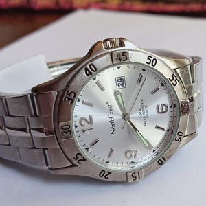 May include: A silver-toned NorthCrest analog wristwatch featuring a metallic link band, a round silver dial with Arabic numerals at 6 and 12, luminous hands, a date window at the 3 o'clock position, and a rotating bezel marked with 5-minute increments.