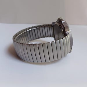 K&ouml;nnte beinhalten: Eine silberfarbene Armbanduhr mit einem dehnbaren Armband. Das Zifferblatt der Uhr ist dunkel, und das Armband besteht aus rechteckigen Metallgliedern. Die Uhr wird auf einer wei&szlig;en Oberfl&auml;che gezeigt.
