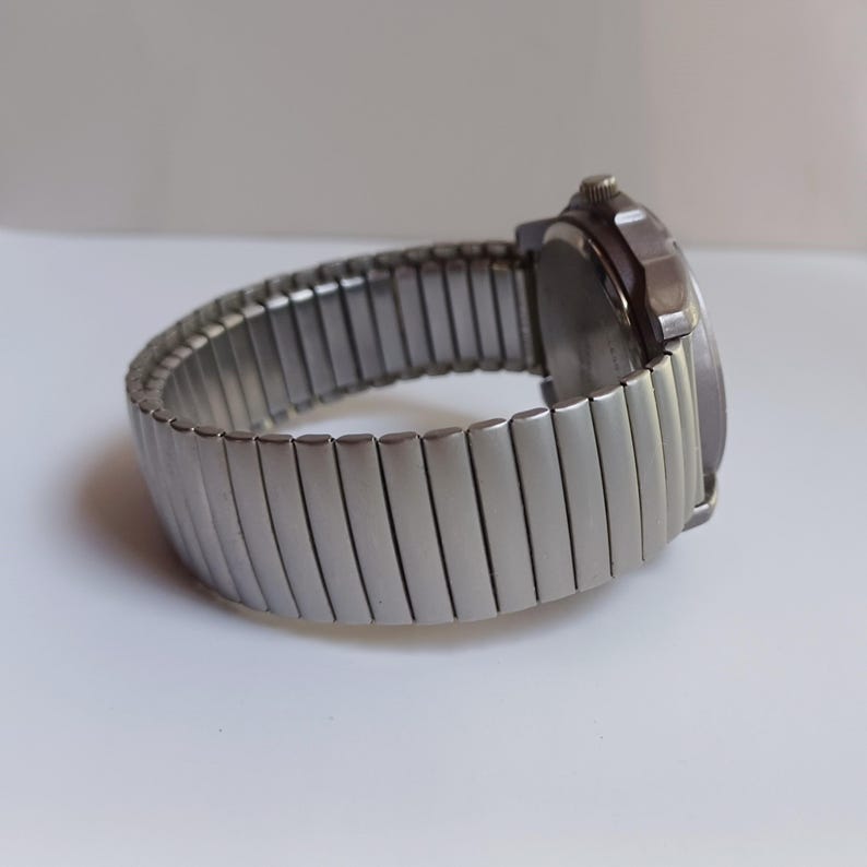 K&ouml;nnte beinhalten: Eine silberfarbene Armbanduhr mit einem dehnbaren Armband. Das Zifferblatt der Uhr ist dunkel, und das Armband besteht aus rechteckigen Metallgliedern. Die Uhr wird auf einer wei&szlig;en Oberfl&auml;che gezeigt.
