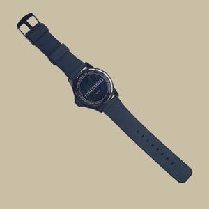 K&ouml;nnte beinhalten: Eine schwarze Armbanduhr mit Silikonarmband und rundem Geh&auml;use. Die Geh&auml;user&uuml;ckseite besteht aus Edelstahl und tr&auml;gt die Aufschrift ROUSSEAU, 15167 und 3 ATM WATER RESISTANT. Die Uhr verf&uuml;gt &uuml;ber einen Dornschlie&szlig;enverschluss und eine matte Oberfl&auml;che.