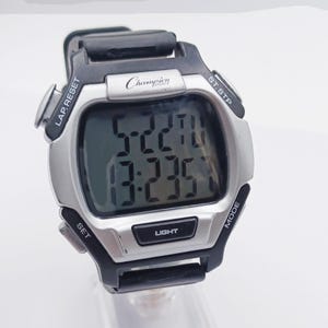 May include: A Champion Sports digital watch with a silver-tone face and black strap. The watch displays the time and has buttons labeled "LAP.RESET", "ST.STP", "MODE", "SET", and "LIGHT". The brand name "Champion" is visible on the face.