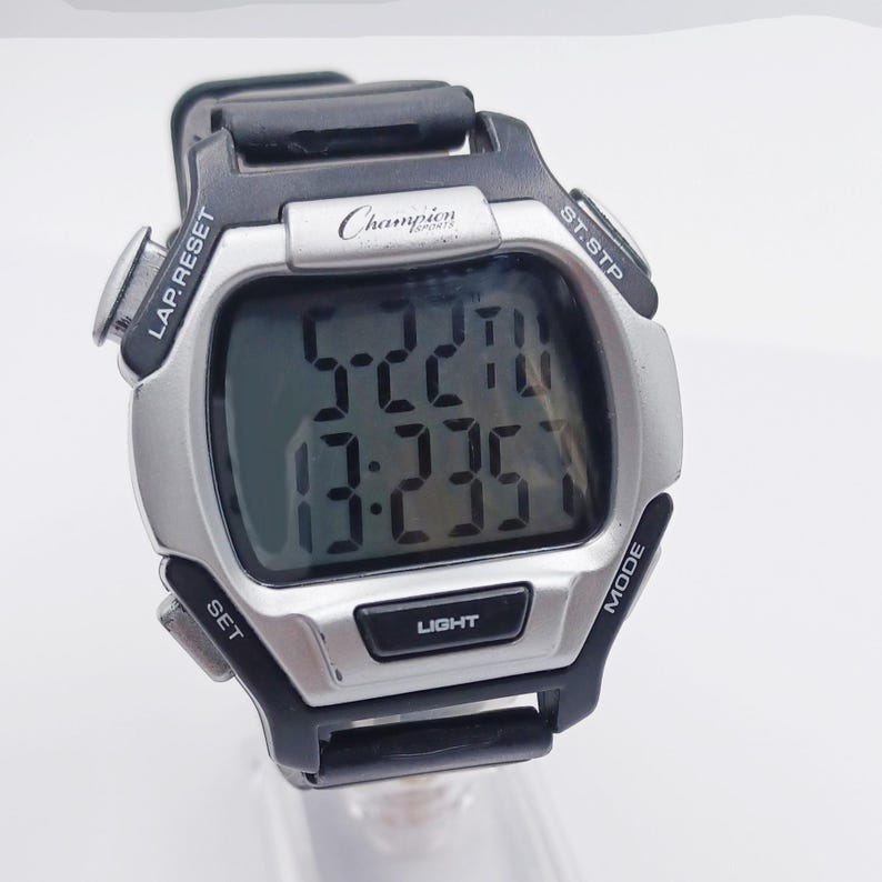 May include: A Champion Sports digital watch with a silver-tone face and black strap. The watch displays the time and has buttons labeled "LAP.RESET", "ST.STP", "MODE", "SET", and "LIGHT". The brand name "Champion" is visible on the face.