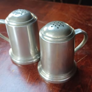 May include: Two silver-colored salt and pepper shakers. Each shaker has a handle and a perforated lid. The shakers are cylindrical with a flared base. The items are made of metal and sit on a wooden surface.
