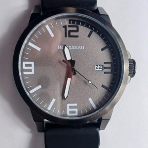 K&ouml;nnte beinhalten: Eine Armbanduhr mit schwarzem Geh&auml;use und schwarzem Silikonarmband. Das Zifferblatt ist strukturiert metallisch grau mit dem Markennamen ROSSEAU, wei&szlig;en Stundenmarkierungen, gro&szlig;en wei&szlig;en Ziffern bei 12, 9 und 6, wei&szlig;en Zeigern und einem Datumsfenster 22.