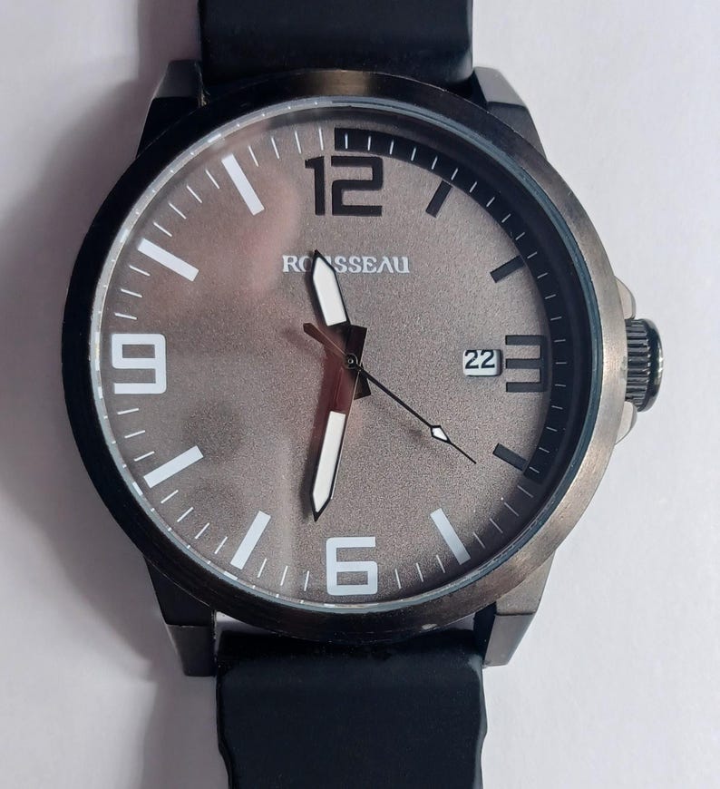 K&ouml;nnte beinhalten: Eine Armbanduhr mit schwarzem Geh&auml;use und schwarzem Silikonarmband. Das Zifferblatt ist strukturiert metallisch grau mit dem Markennamen ROSSEAU, wei&szlig;en Stundenmarkierungen, gro&szlig;en wei&szlig;en Ziffern bei 12, 9 und 6, wei&szlig;en Zeigern und einem Datumsfenster 22.