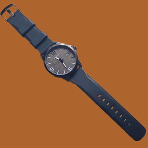 K&ouml;nnte beinhalten: Eine dunkelblaue Armbanduhr mit rundem Geh&auml;use, dunkelgrauem Zifferblatt, wei&szlig;en Stundenmarkierungen und gro&szlig;en wei&szlig;en Ziffern auf den 12-, 6- und 9-Uhr-Positionen. Die Uhr verf&uuml;gt &uuml;ber ein Datumsfenster und ein flexibles dunkelblaues Silikonarmband.