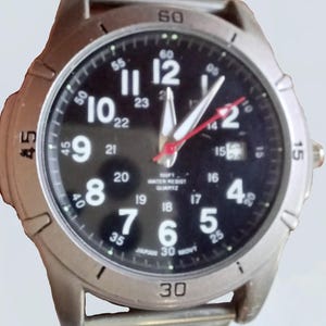 K&ouml;nnte beinhalten: Eine silberfarbene Armbanduhr mit schwarzem Zifferblatt. Die Uhr hat wei&szlig;e Ziffern, wei&szlig;e Zeiger und einen roten Sekundenzeiger. Auf dem Zifferblatt steht "100FT WATER RESIST QUARTZ" und "JAPAN 30 MOVT".