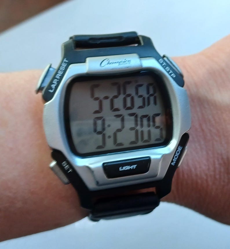 May include: A Champion digital watch with a silver face and black accents. The watch displays the time and has buttons labeled "LAR RESET", "BT.STP", "SET", "MODE", and "LIGHT". The watch is on a wrist.