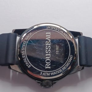 K&ouml;nnte beinhalten: Die R&uuml;ckseite einer Rousseau-Armbanduhr mit dunkelblauem Silikonarmband. Der kreisf&ouml;rmige Geh&auml;useboden aus Edelstahl tr&auml;gt die eingravierten Texte ROUSSEAU, 15167, STAINLESS STEEL BACK, JAPAN MOVEMENT und 3 ATM WATER RESISTANT auf der Oberfl&auml;che.