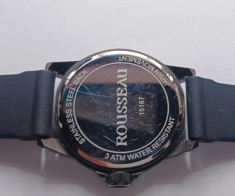 K&ouml;nnte beinhalten: Die R&uuml;ckseite einer Rousseau-Armbanduhr mit dunkelblauem Silikonarmband. Der kreisf&ouml;rmige Geh&auml;useboden aus Edelstahl tr&auml;gt die eingravierten Texte ROUSSEAU, 15167, STAINLESS STEEL BACK, JAPAN MOVEMENT und 3 ATM WATER RESISTANT auf der Oberfl&auml;che.