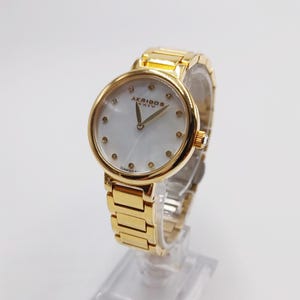 May include: Gold-toned Akribos XXIV watch with a white mother-of-pearl face. The watch features gold hands, diamond hour markers, and a gold link bracelet. The watch is displayed on a clear stand.
