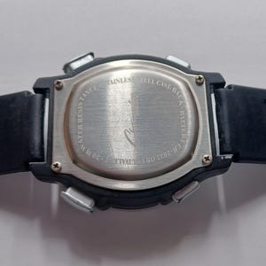 May include: Close-up of the back of a black digital watch. The watch has a stainless steel back with text that reads "Stainless Steel Case Back Battery" and "Water Resist 30 M". The watch band is black.