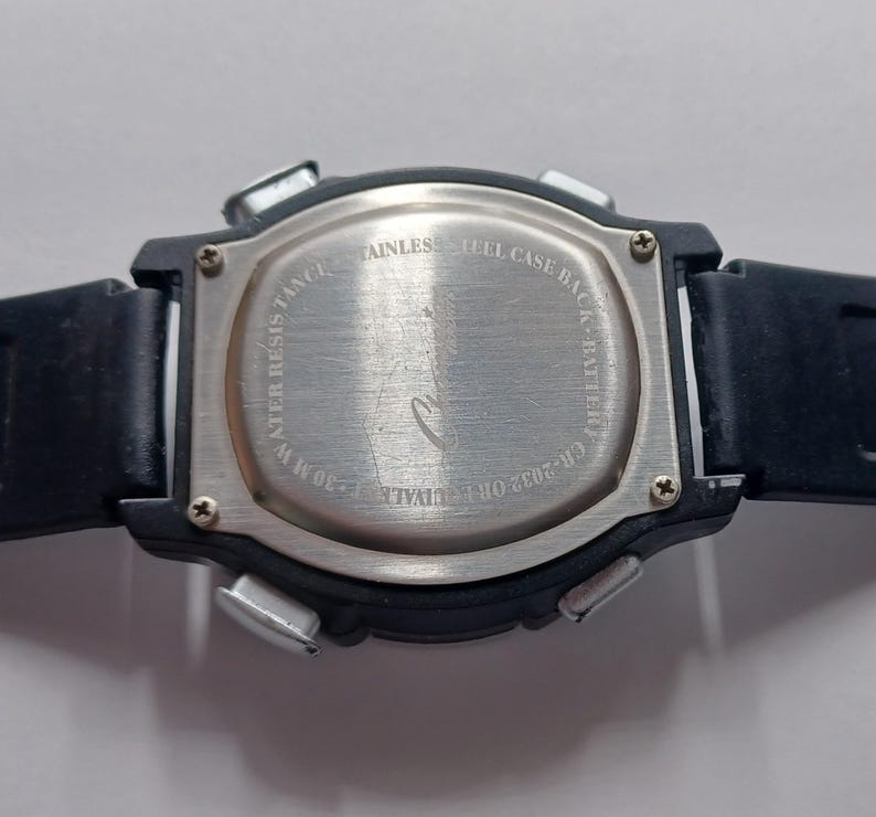 May include: Close-up of the back of a black digital watch. The watch has a stainless steel back with text that reads "Stainless Steel Case Back Battery" and "Water Resist 30 M". The watch band is black.
