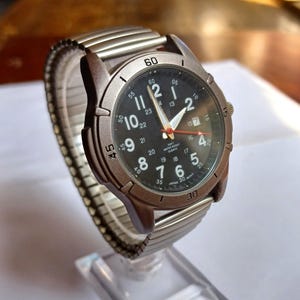 K&ouml;nnte beinhalten: Armbanduhr mit schwarzem Zifferblatt, wei&szlig;en Ziffern und Zeigern sowie Datumsanzeige. Die Uhr hat eine braune L&uuml;nette und ein silberfarbenes, dehnbares Metallarmband. Die Uhr wird auf einem transparenten St&auml;nder pr&auml;sentiert. Der Zifferblattdurchmesser betr&auml;gt ca. 4 cm.