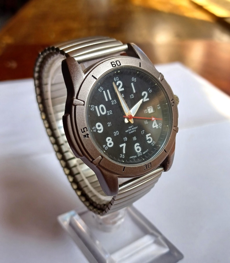 K&ouml;nnte beinhalten: Armbanduhr mit schwarzem Zifferblatt, wei&szlig;en Ziffern und Zeigern sowie Datumsanzeige. Die Uhr hat eine braune L&uuml;nette und ein silberfarbenes, dehnbares Metallarmband. Die Uhr wird auf einem transparenten St&auml;nder pr&auml;sentiert. Der Zifferblattdurchmesser betr&auml;gt ca. 4 cm.