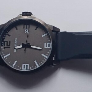 K&ouml;nnte beinhalten: Eine Armbanduhr mit dunkelgrauem, rundem Zifferblatt, wei&szlig;en Stundenmarkierungen und gro&szlig;en wei&szlig;en Ziffern auf den 6-, 9- und 12-Uhr-Positionen. Die Uhr verf&uuml;gt &uuml;ber ein Datumsfenster auf der 3-Uhr-Position, schwarze Zeiger und ein dunkelblaues Silikonarmband.