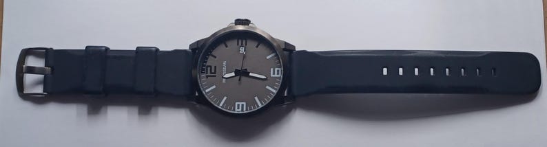 K&ouml;nnte beinhalten: Eine Armbanduhr mit dunkelgrauem, rundem Zifferblatt, wei&szlig;en Stundenmarkierungen und gro&szlig;en wei&szlig;en Ziffern auf den 6-, 9- und 12-Uhr-Positionen. Die Uhr verf&uuml;gt &uuml;ber ein Datumsfenster auf der 3-Uhr-Position, schwarze Zeiger und ein dunkelblaues Silikonarmband.