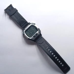 May include: A digital wristwatch with a black rubber strap and a silver-colored face. The watch displays the time and date on a digital screen. The watch has buttons on the side and a buckle closure. The watch is lying on a white surface.