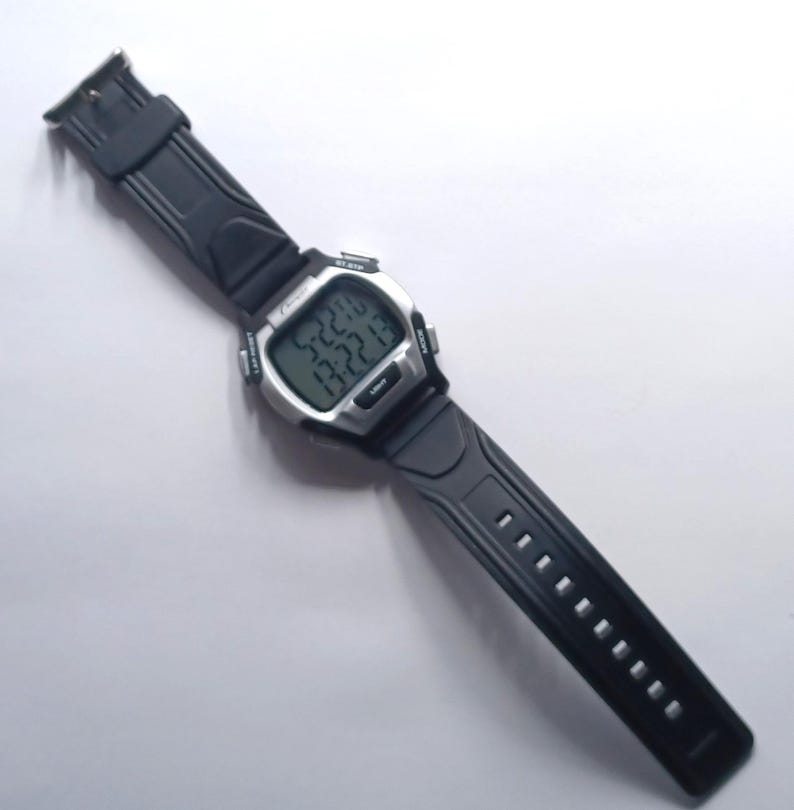 May include: A digital wristwatch with a black rubber strap and a silver-colored face. The watch displays the time and date on a digital screen. The watch has buttons on the side and a buckle closure. The watch is lying on a white surface.