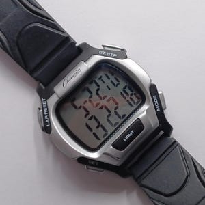 May include: A Champion digital watch with a silver-tone case and black rubber strap. The watch face displays the time and other functions. The watch has buttons for light, mode, set, and lap reset. The watch is designed for sports and everyday wear.