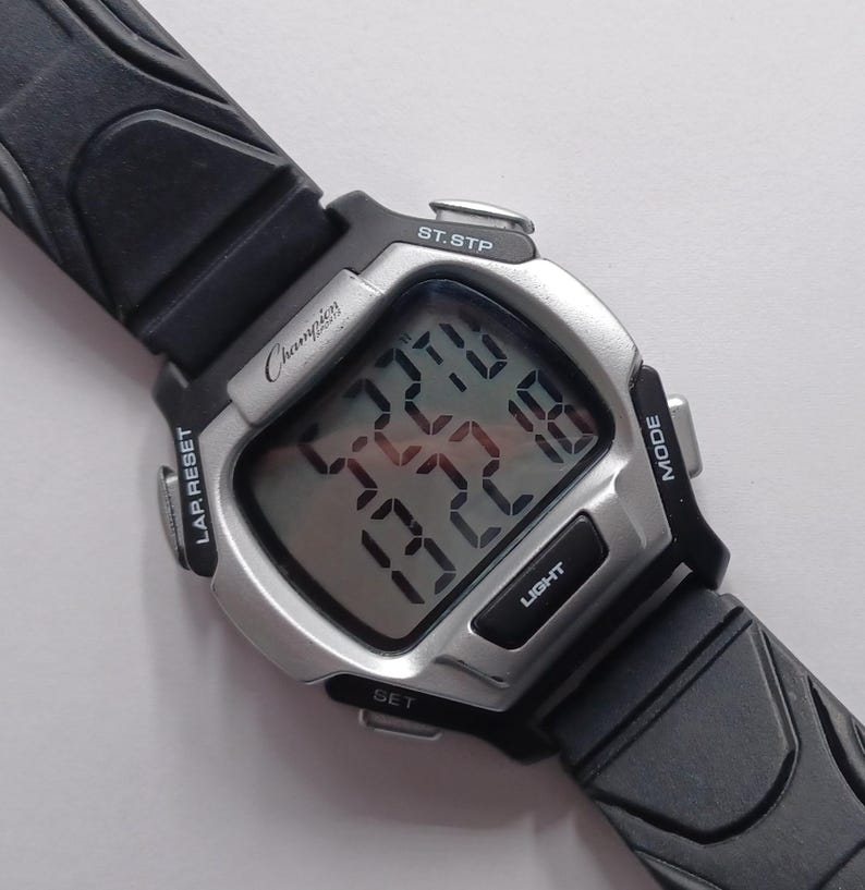 May include: A Champion digital watch with a silver-tone case and black rubber strap. The watch face displays the time and other functions. The watch has buttons for light, mode, set, and lap reset. The watch is designed for sports and everyday wear.