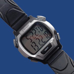 May include: A Champion digital wristwatch with a silver-tone case and black rubber strap. The watch face displays the time and other functions. Features include a light button, mode button, and lap reset.