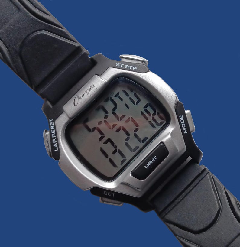 May include: A Champion digital wristwatch with a silver-tone case and black rubber strap. The watch face displays the time and other functions. Features include a light button, mode button, and lap reset.