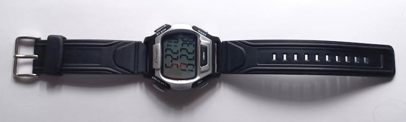 May include: A digital wristwatch with a black rubber strap and a silver-colored face. The watch displays the time and date on a digital screen. The strap has a buckle closure and multiple adjustment holes. The watch is designed for everyday wear.